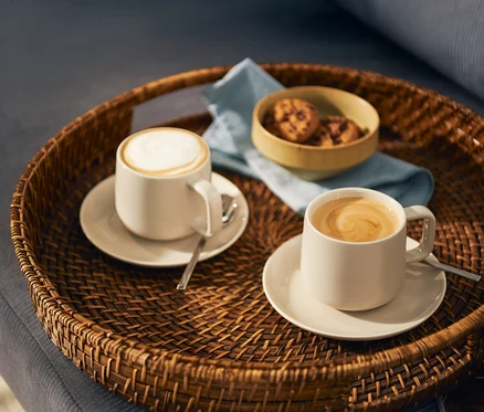 Dva světlé šálky kávy, jeden s mléčnou pěnou a druhý s latte art spirálou, stojí na odpovídajících podšálcích s lžičkami na kulatém proutěném podnosu. Na podnosu je také malá miska s sušenkami a světle modrý ubrousek.