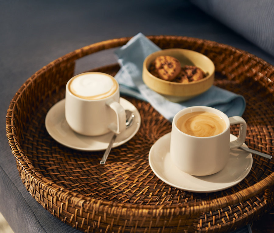 Dva světlé šálky kávy, jeden s mléčnou pěnou a druhý s latte art spirálou, stojí na odpovídajících podšálcích s lžičkami na kulatém proutěném podnosu. Na podnosu je také malá miska s sušenkami a světle modrý ubrousek.