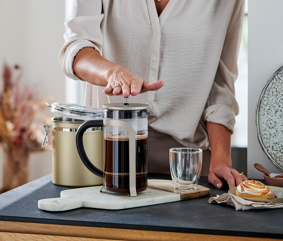 Osoba stlačuje píst konvičky french press (800 ml, 6 šálků, šampaň) naplněné kávou. Vedle je sklenice a zákusek.