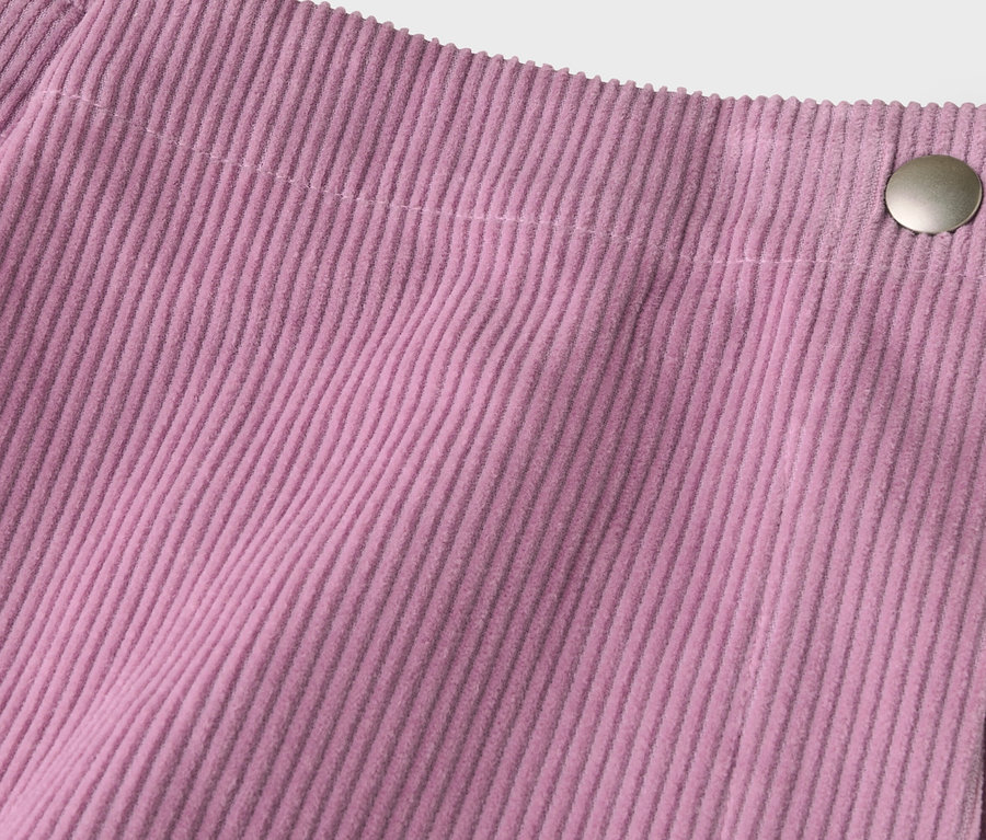 Detail růžové manšestrové látky s vertikálními žebry a stříbrným knoflíkem vpravo nahoře, který zajišťuje překrývající se okraj.