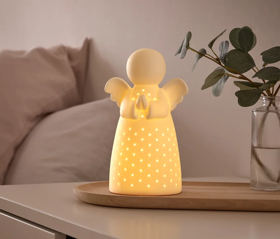 Svítící bílá keramická lampa ve tvaru anděla s malými perforacemi stojí na dřevěném podnosu na nočním stolku, vedle vázy se zelenou rostlinou. V pozadí je postel se světle růžovými polštáři.