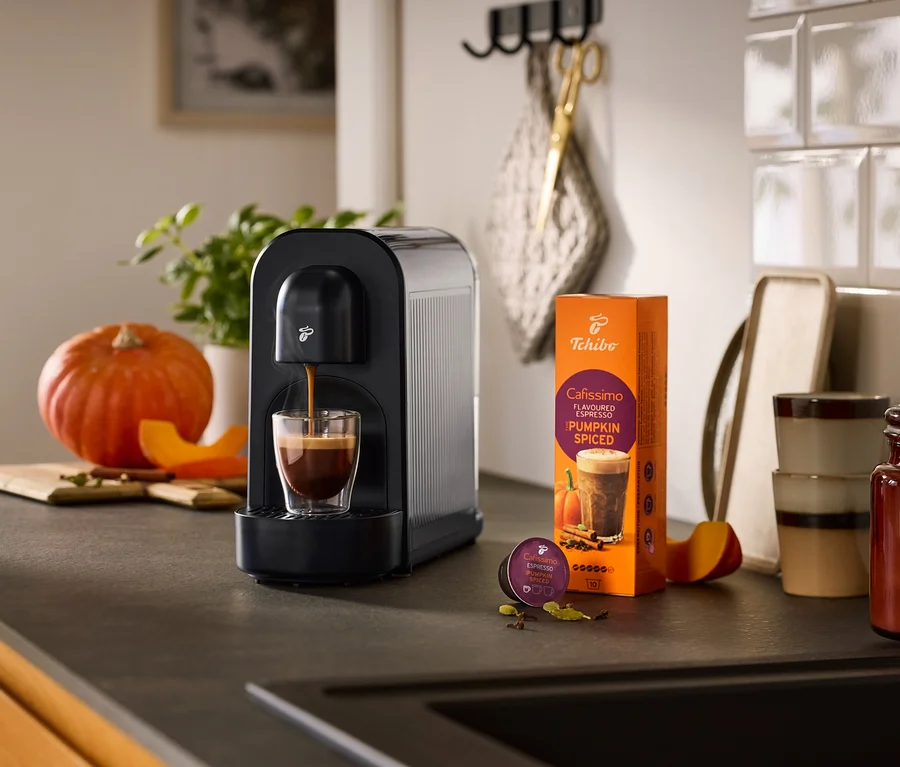 Černý kávovar Cafissimo now připravuje espresso do sklenice, vedle něj balení kávy Cafissimo Pumpkin Spiced a kapsle, obklopené dýní a kořením na kuchyňské lince.