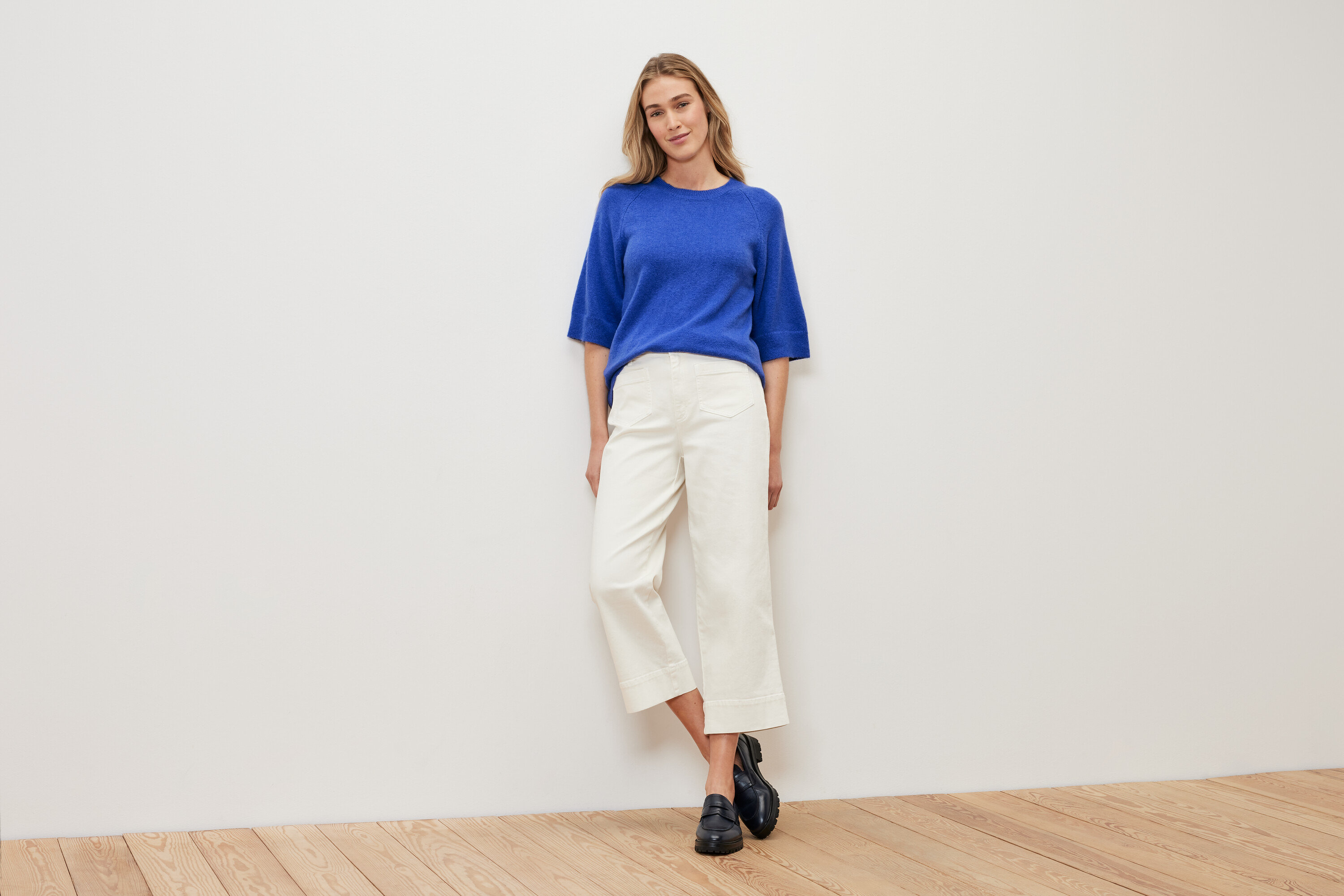 Žena stojící před bílou zdí má na sobě pletený svetr kobaltové modré barvy, bílé kalhoty a černé boty.