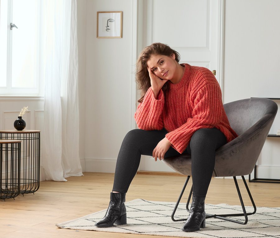 Žena sedí v křesle a má na sobě huňatý svetr, džínové legíny a černé kožené kotníkové boty.