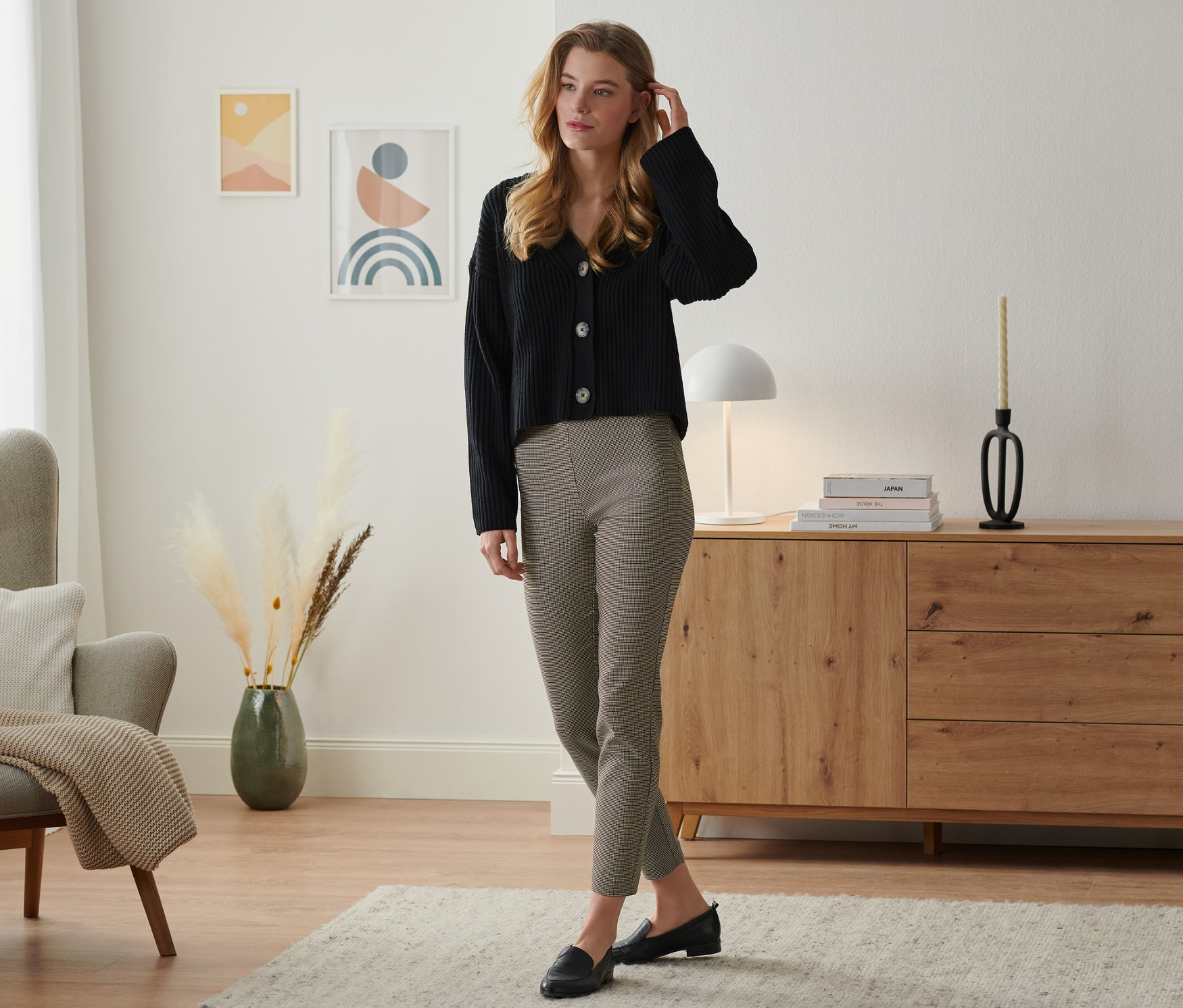 Žena stojí v obývacím pokoji a má na sobě černé sako a strečové kalhoty.