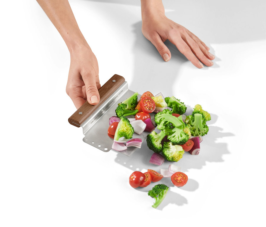 Dvě ruce drží kráječ na těsto 2 v 1 s nakrájenou brokolicí, rajčaty a cibulí na bílém pozadí.