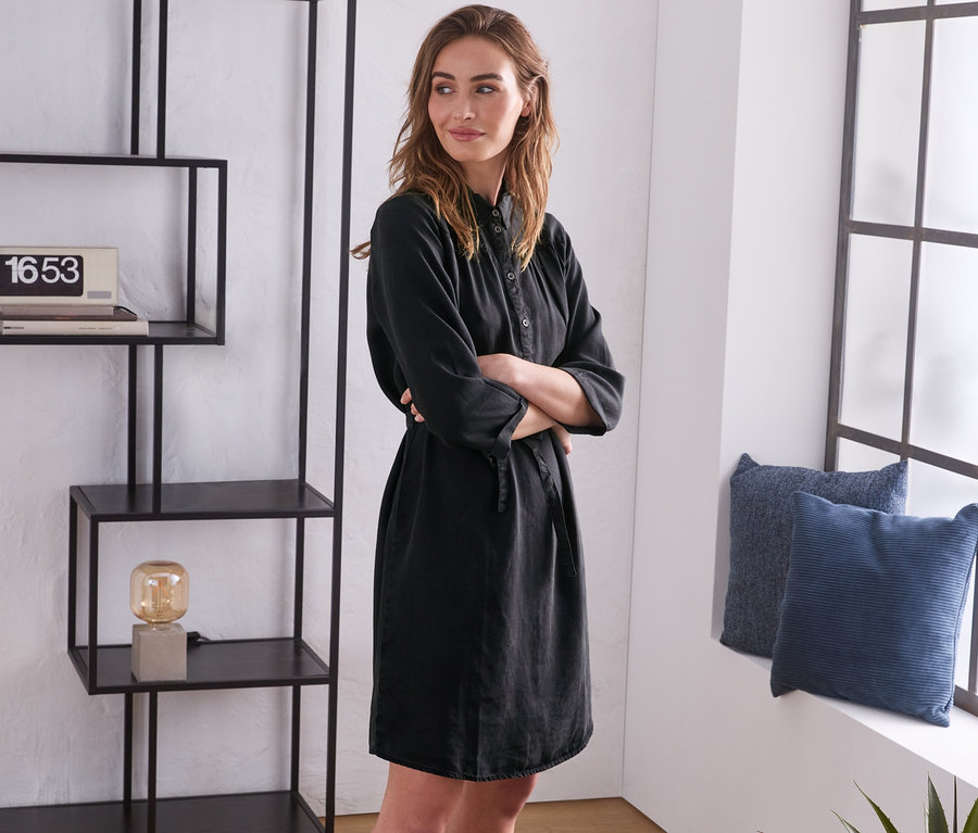 Žena stojí a má zkřížené ruce na sobě denimové šaty.