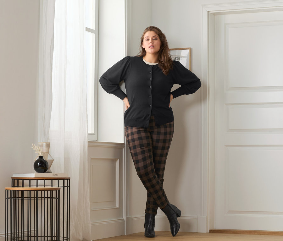 Žena pózuje v kabátku z jemného úpletu s objemnými rukávy, kostkovaných strečových kalhotách a kožených kotníkových botách.