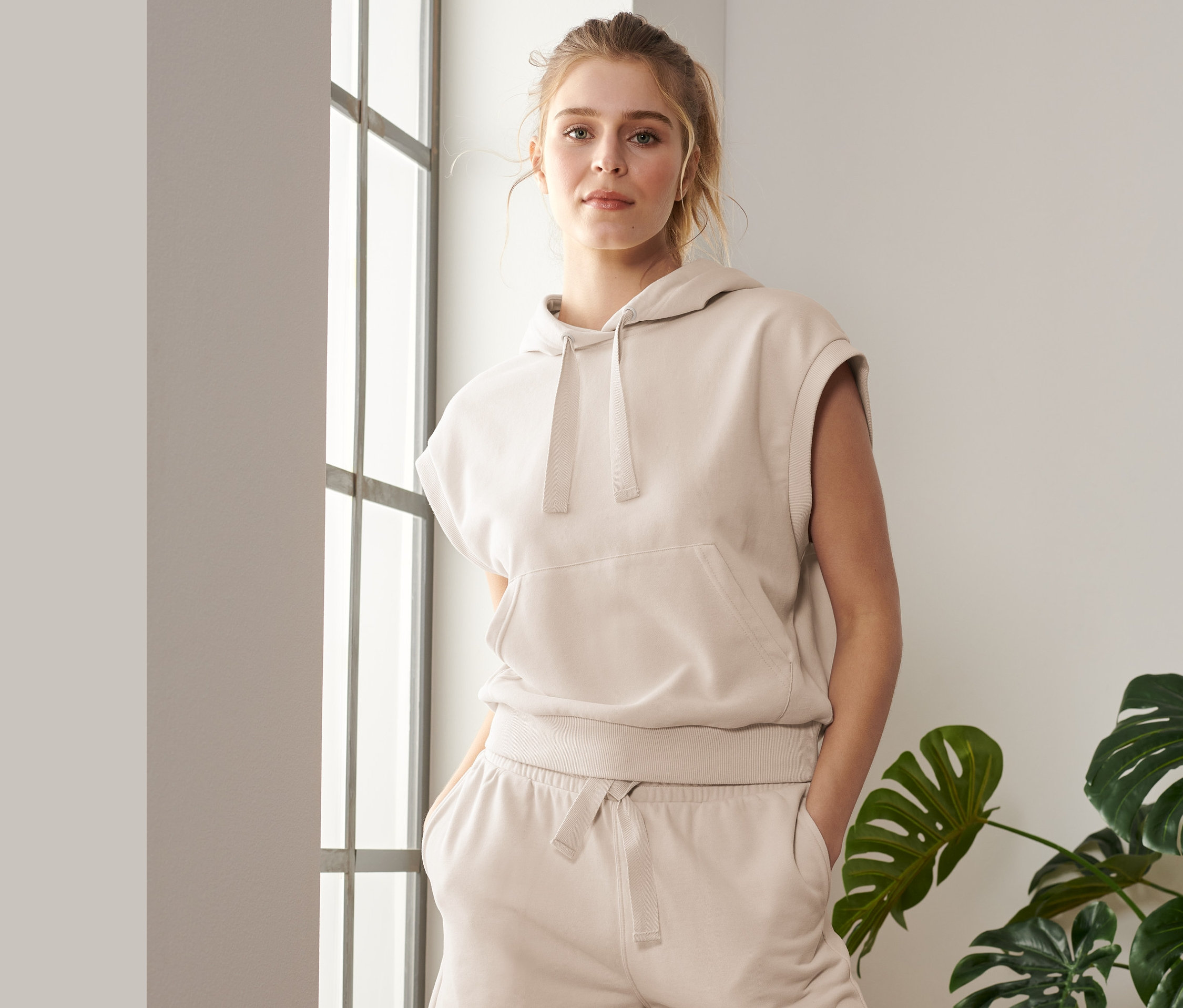 Žena pózuje u okna ve sportovní mikině bez rukávů a šortkách.