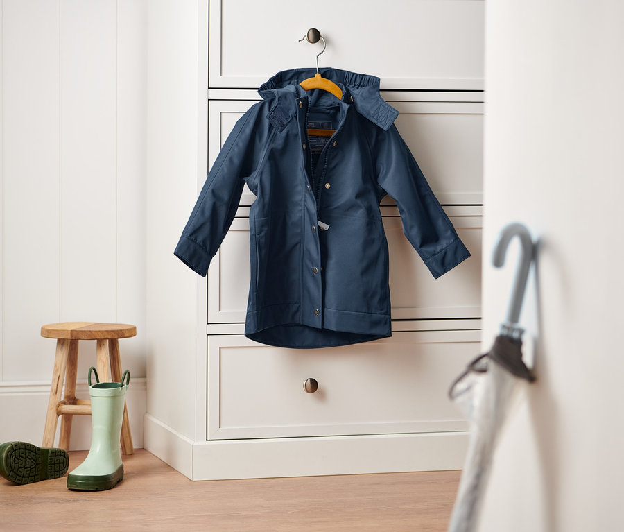 Tmavě modrá bunda do deště visí na bílé komodě. Na zemi stojí dřevěná stolička a pár zelených holínek.