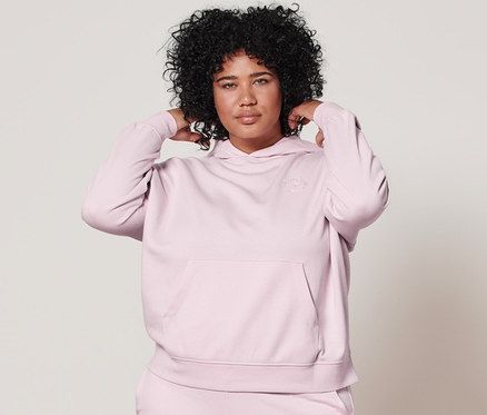 Žena pózuje v růžové mikině s kapucí a teplákách.