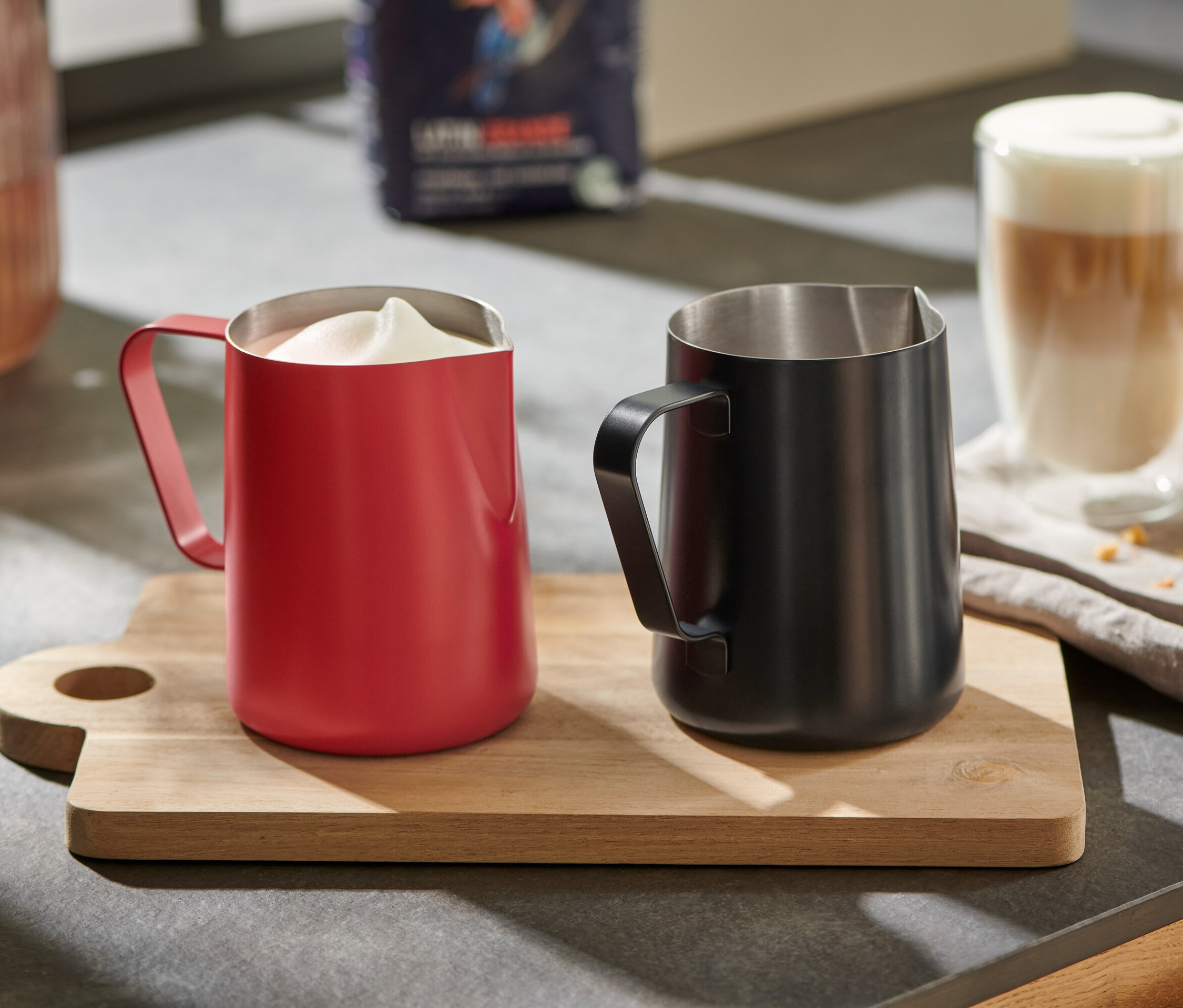 Červená konvička na mléko s napěněným mlékem a černá konvička na mléko na dřevěném prkénku.