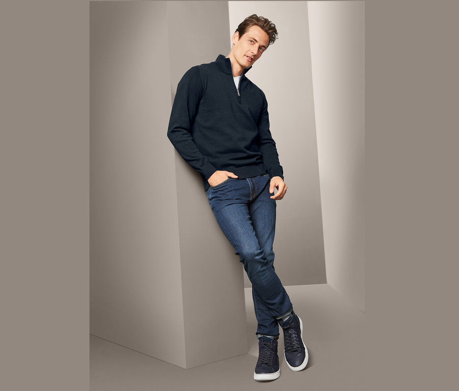 Muž opřený o zeď má na sobě tmavě modrý svetr troyer z hrubé pleteniny, džíny a tenisky.