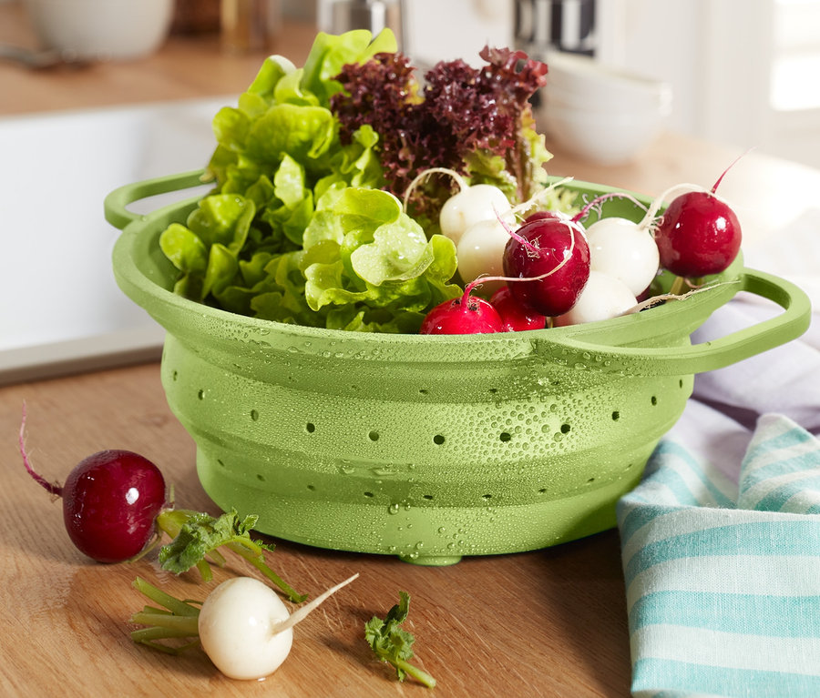Zelený skládací cedník plný salátu a ředkviček stojí na dřevěné kuchyňské desce.