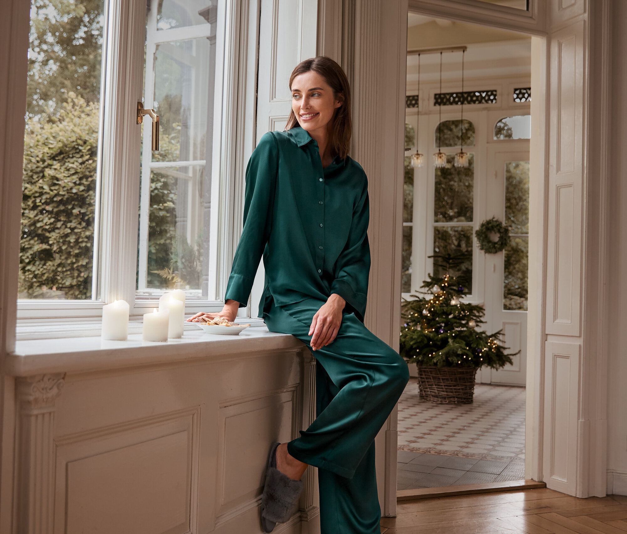 Žena sedí na parapetu v zeleném hedvábném pyžamu a plyšových pantoflích.