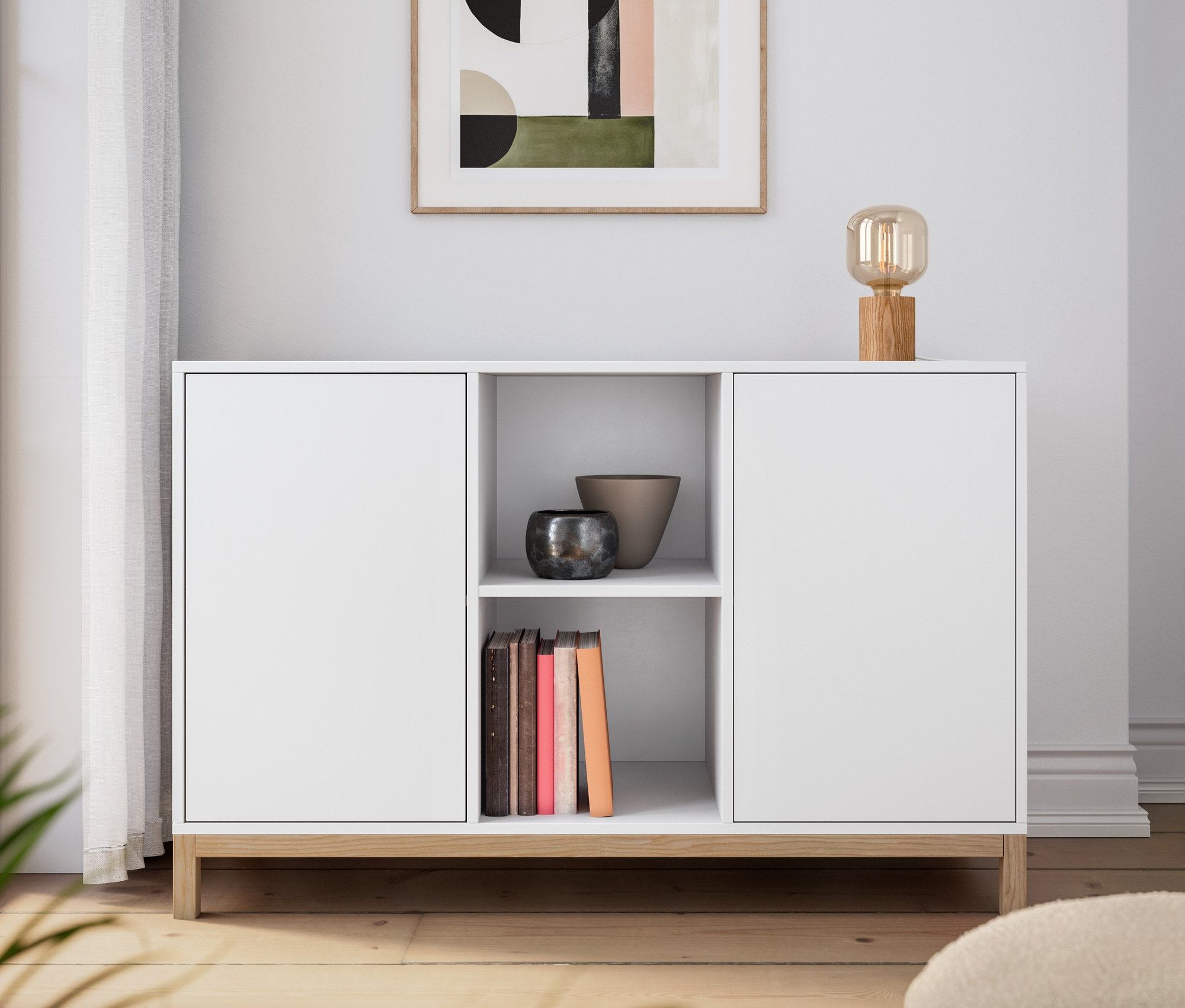 Bílá komoda se stolní lampou ze dřeva. Na zdi visí obraz.