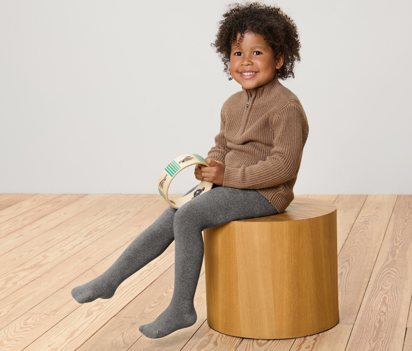 Usmívající se dítě sedí na dřevěné stoličce a drží tamburínu. Má na sobě hnědý svetr a šedé melírované dětské termopunčocháče, 2 ks.