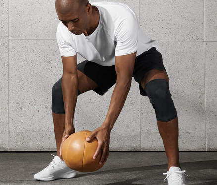 Muž v bílém tričku a černých šortkách drží medicinbal a má na sobě fitness nákolenky, 2 ks.