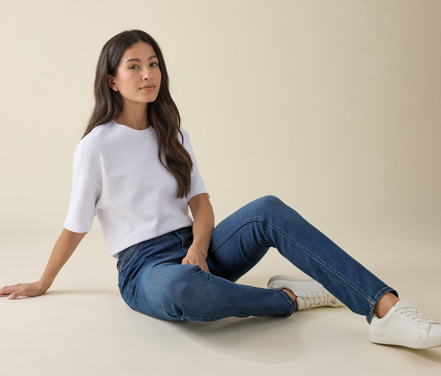 Žena sedí na zemi v džínách slim střihu »Emma«, mid blue denim, bílém tričku a teniskách.