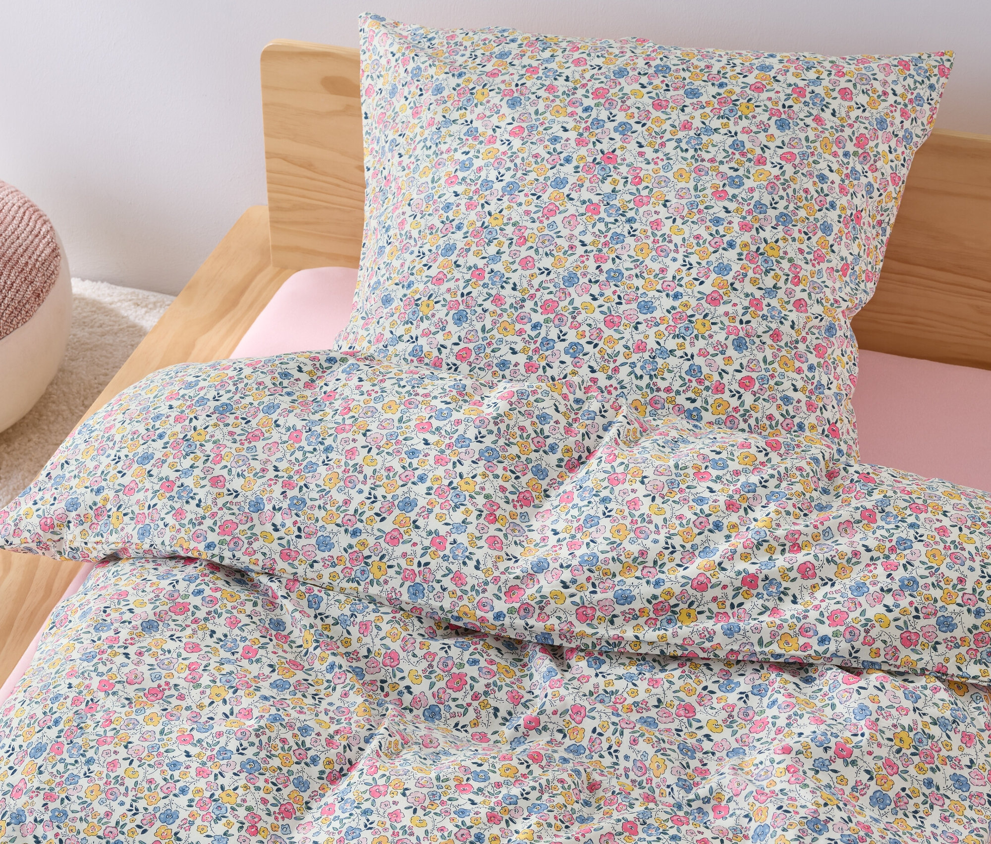 Na posteli je dětské povlečení renforcé standardní velikosti s květinovým vzorem a vedle ní stojí růžový puf.