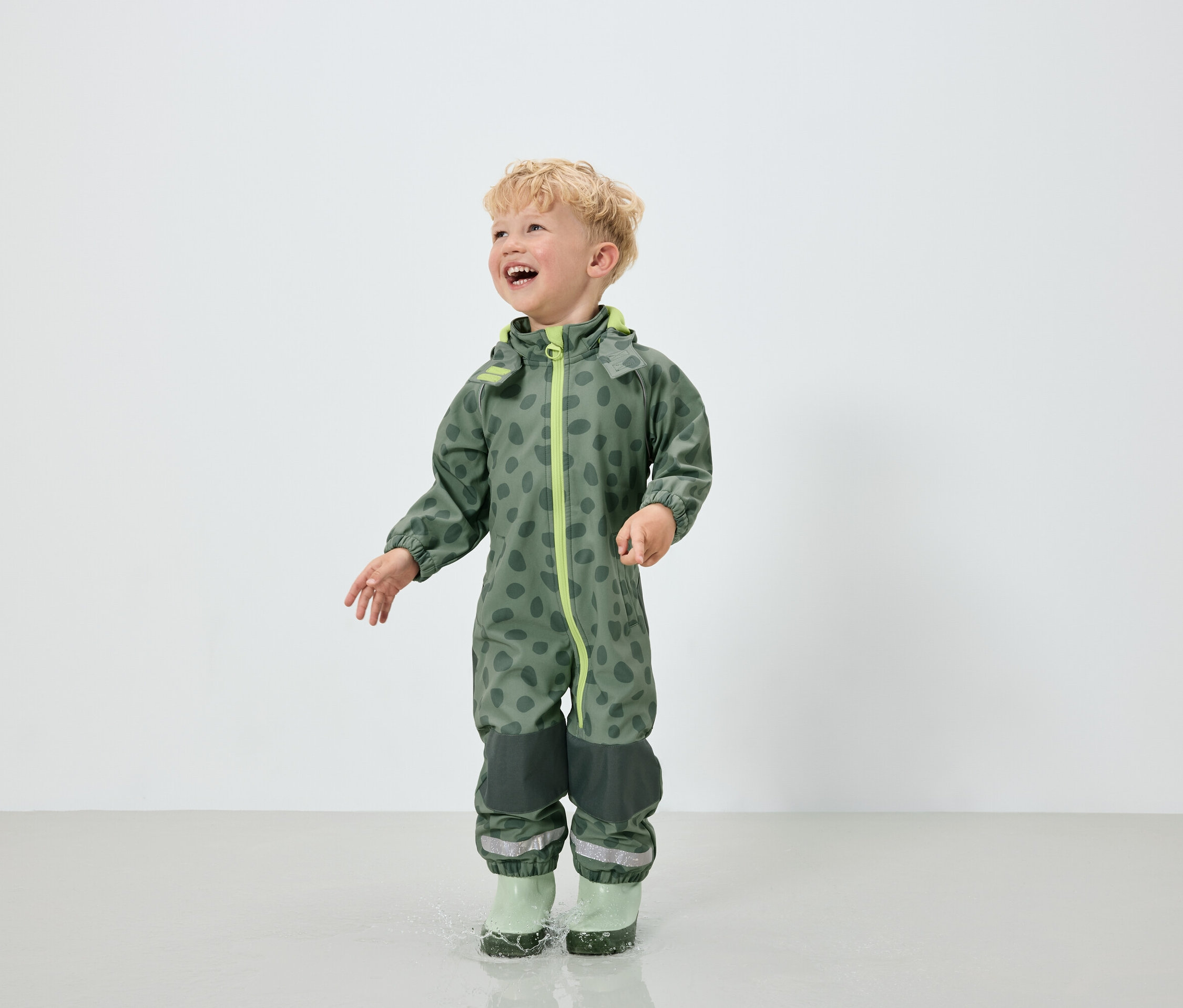 Chlapec v zelené kombinéze se vzorem skvrn a zelených gumákách skáče v kaluži.
