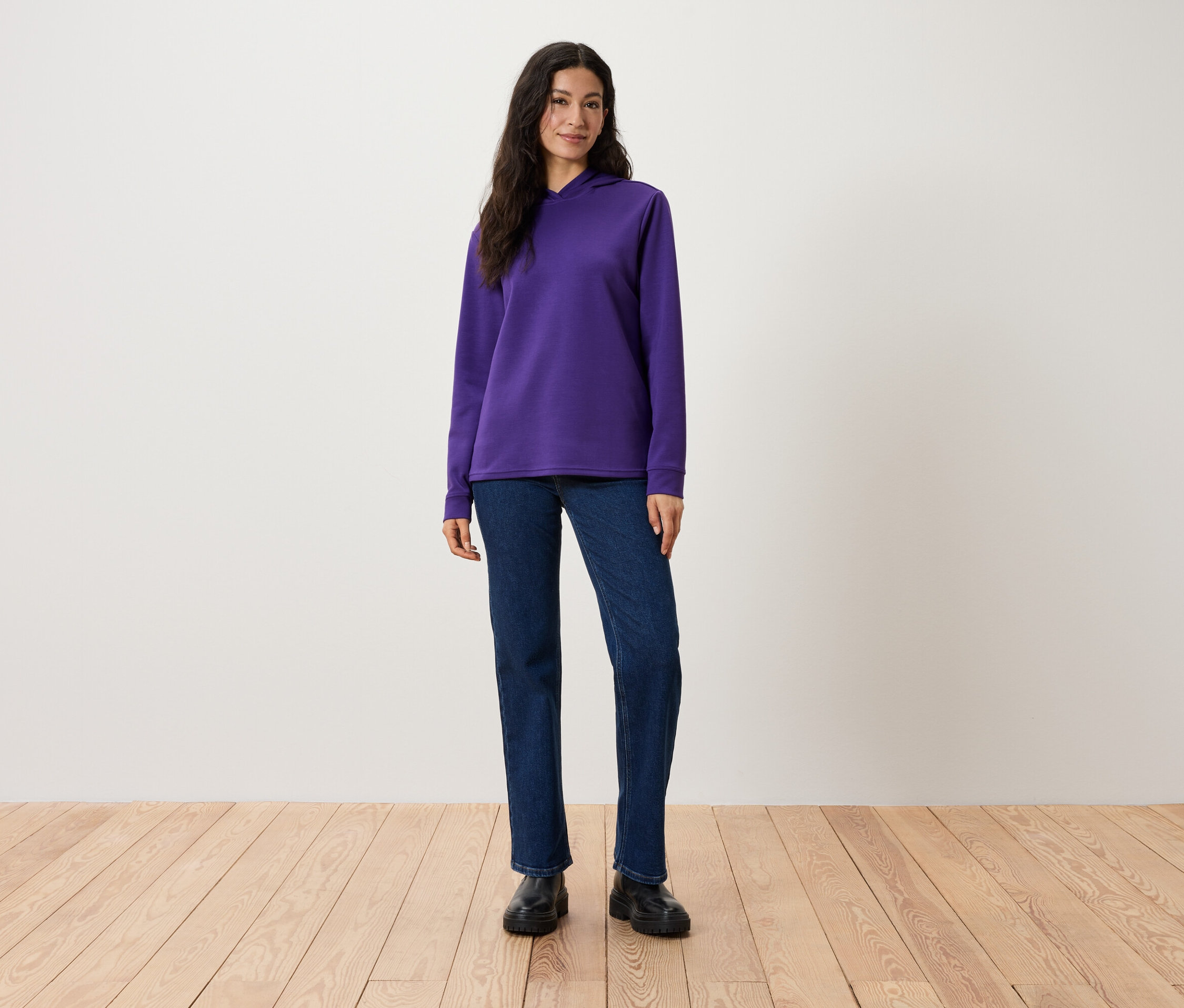Žena stojí a má na sobě fialovou mikinu s kapucí, džíny a černé boty.