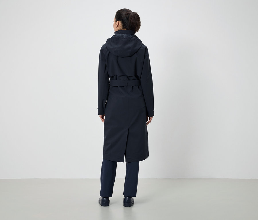 Zezadu je vidět žena s culíkem, která má na sobě tmavě modrý kabát s kapucí a pásek a tmavě modré kalhoty.