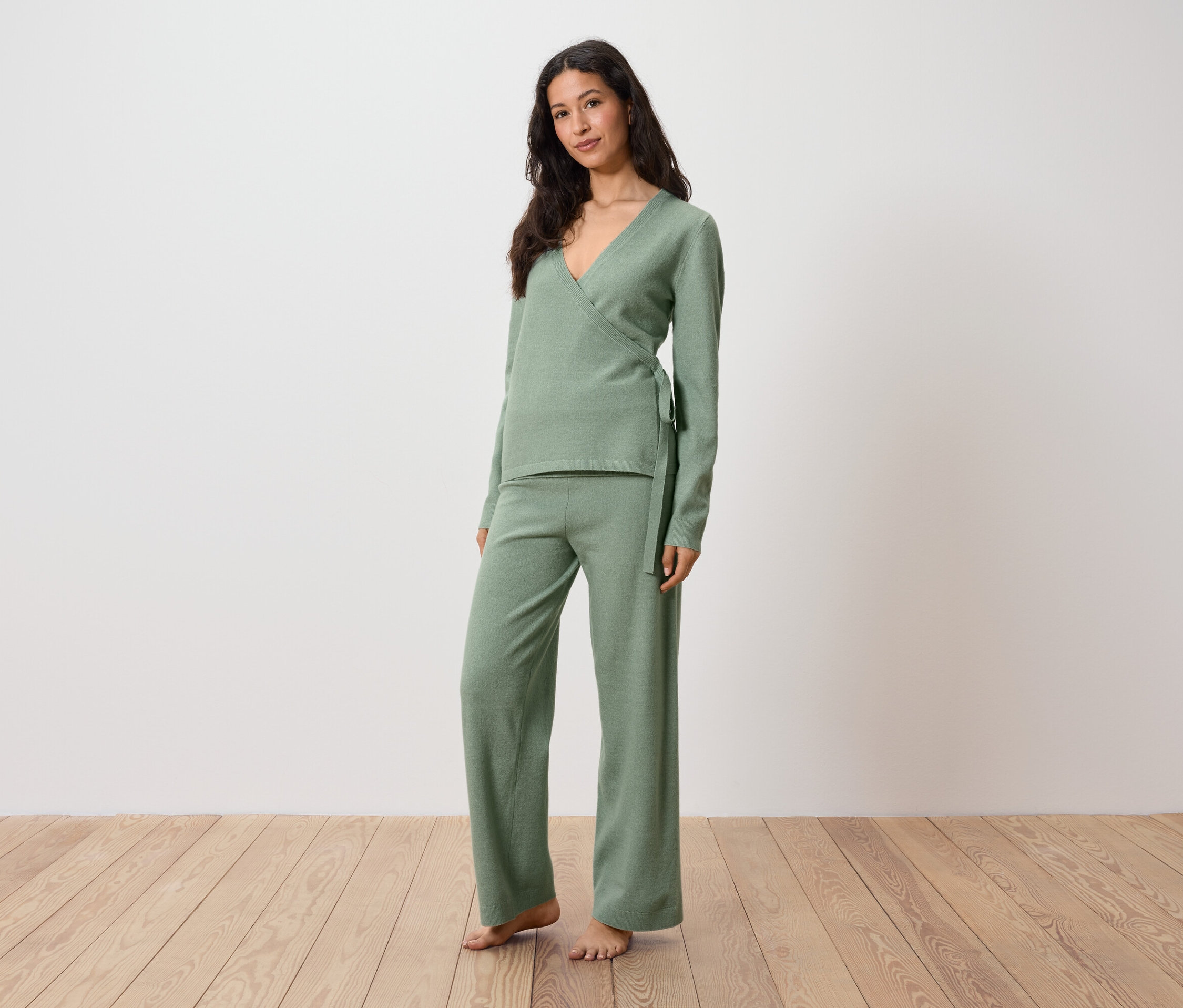 Žena v zeleném svetru a kalhotách stojí na dřevěné podlaze.