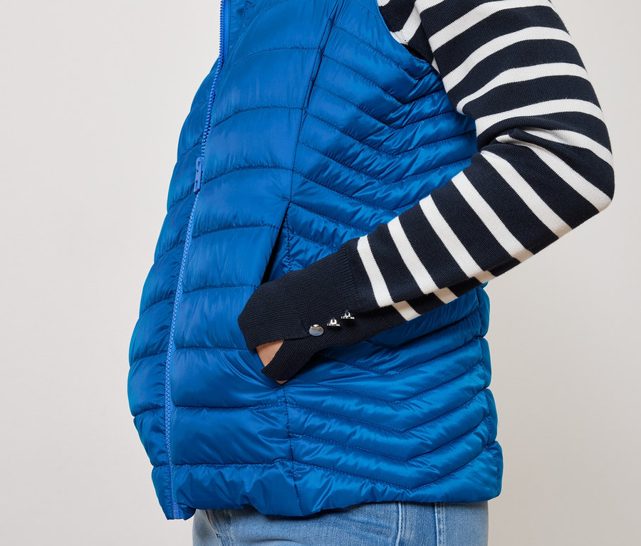 Člověk má ruku v kapse modré prošívané vesty a na sobě tričko s modrobílými pruhy.