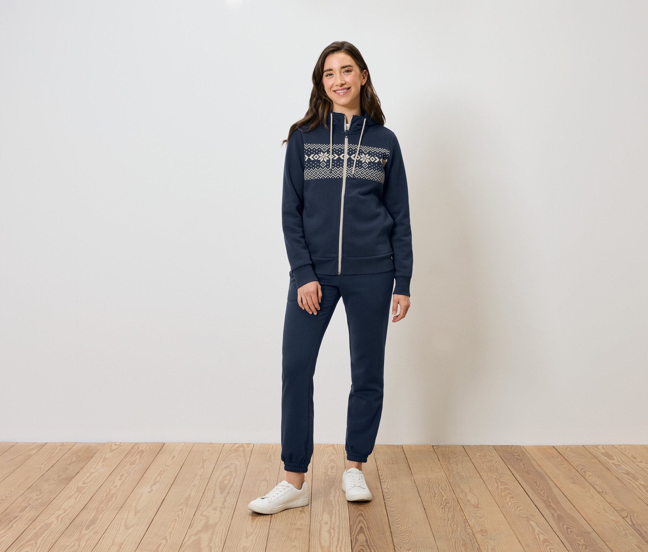 Žena stojí na dřevěné podlaze a má na sobě tmavě modrou mikinu na zip a tepláky a bílé tenisky.