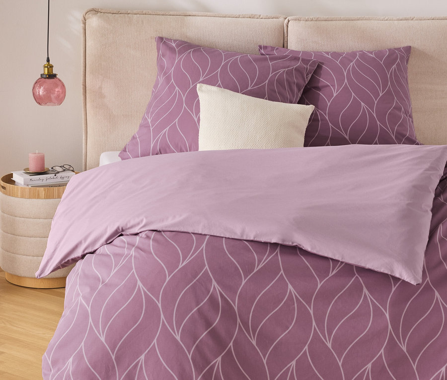 Fialové ložní prádlo s bílým vzorem listů na posteli s béžovým čelem. Noční stolek s lampou stojí vedle postele.