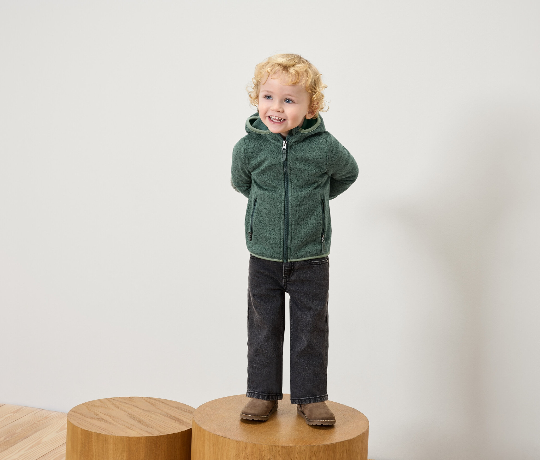 Chlapec s blonďavými kudrnatými vlasy stojí na dřevěném podstavci a má na sobě zelenou mikinu s kapucí, černé džíny a hnědé boty.