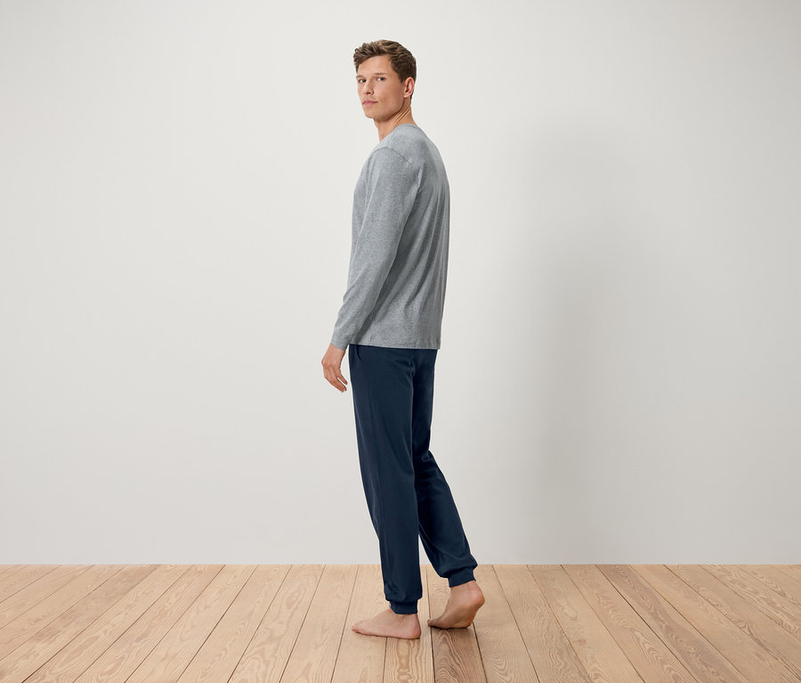 Muž v šedém tričku s dlouhým rukávem a tmavě modrých kalhotách stojí bosý na dřevěné podlaze a dívá se přes rameno.