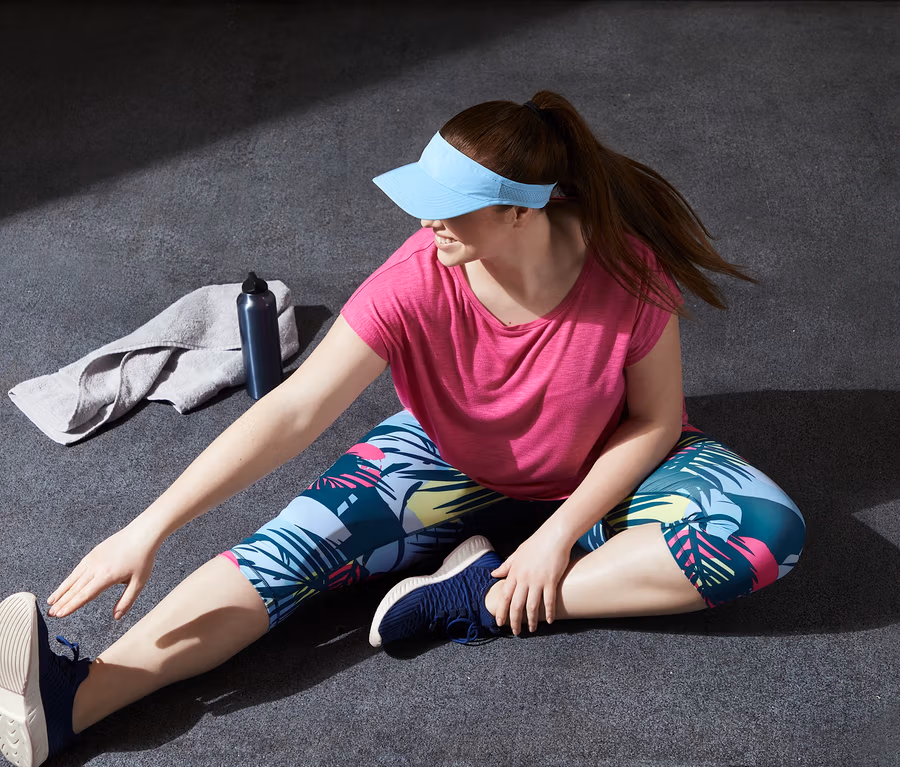 Žena sedí na zemi a protahuje se, má na sobě růžové sportovní tričko a 3/4 sportovní legíny.