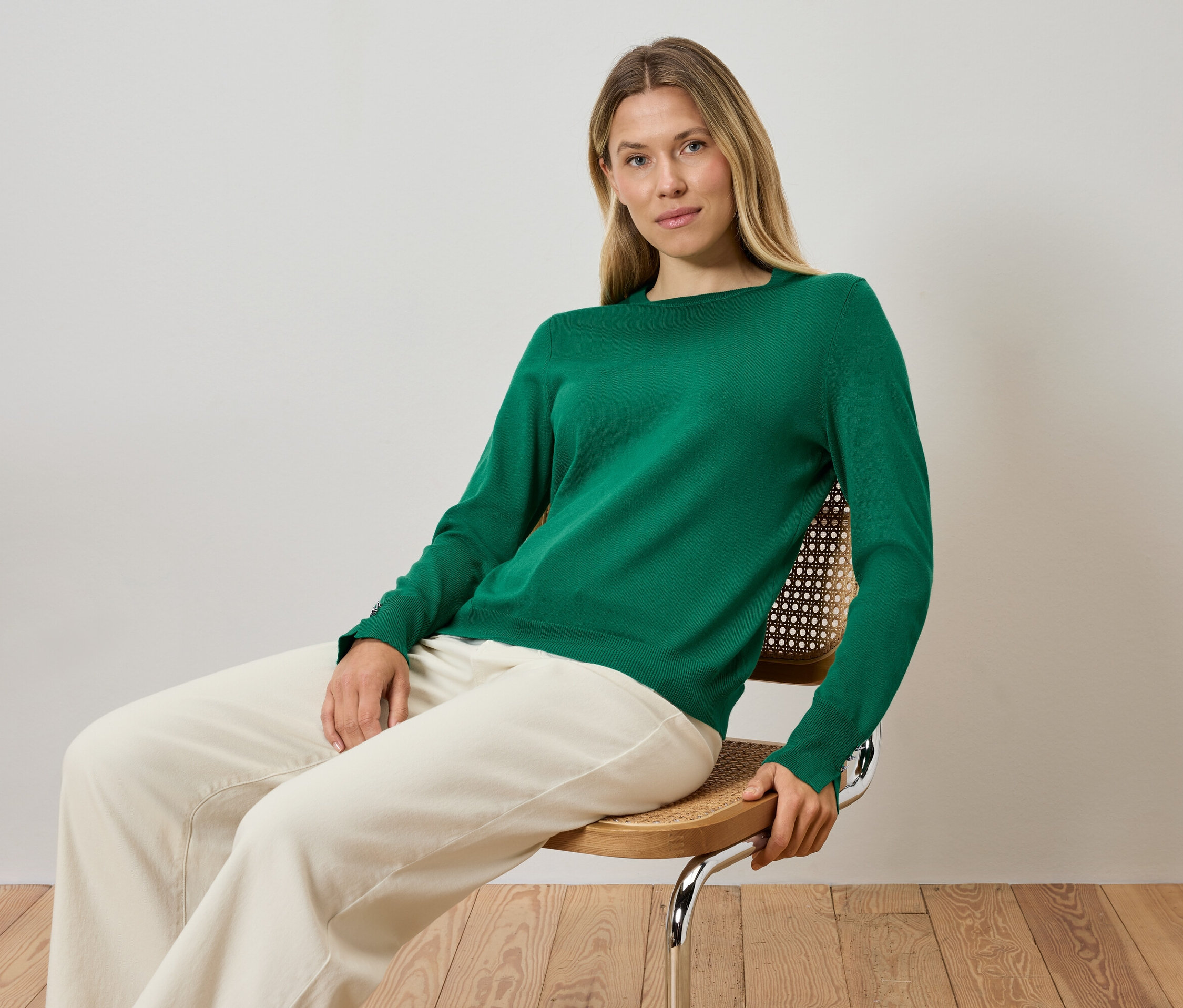 Blondýna sedí na židli a má na sobě zelený svetr a krémové kalhoty.