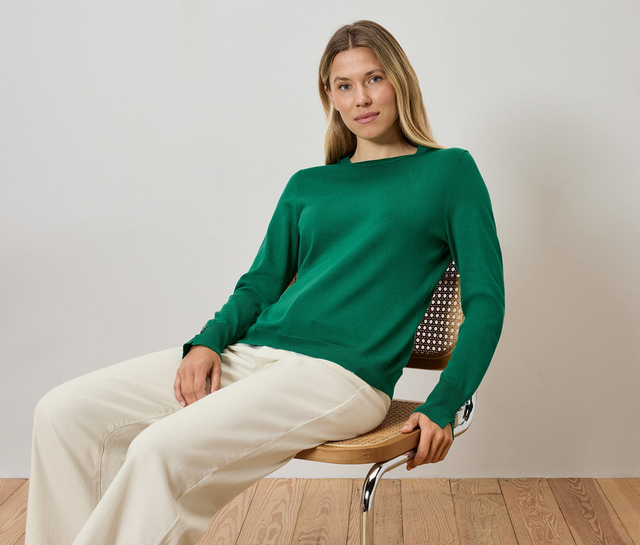 Blondýna sedí na židli a má na sobě zelený svetr a krémové kalhoty.