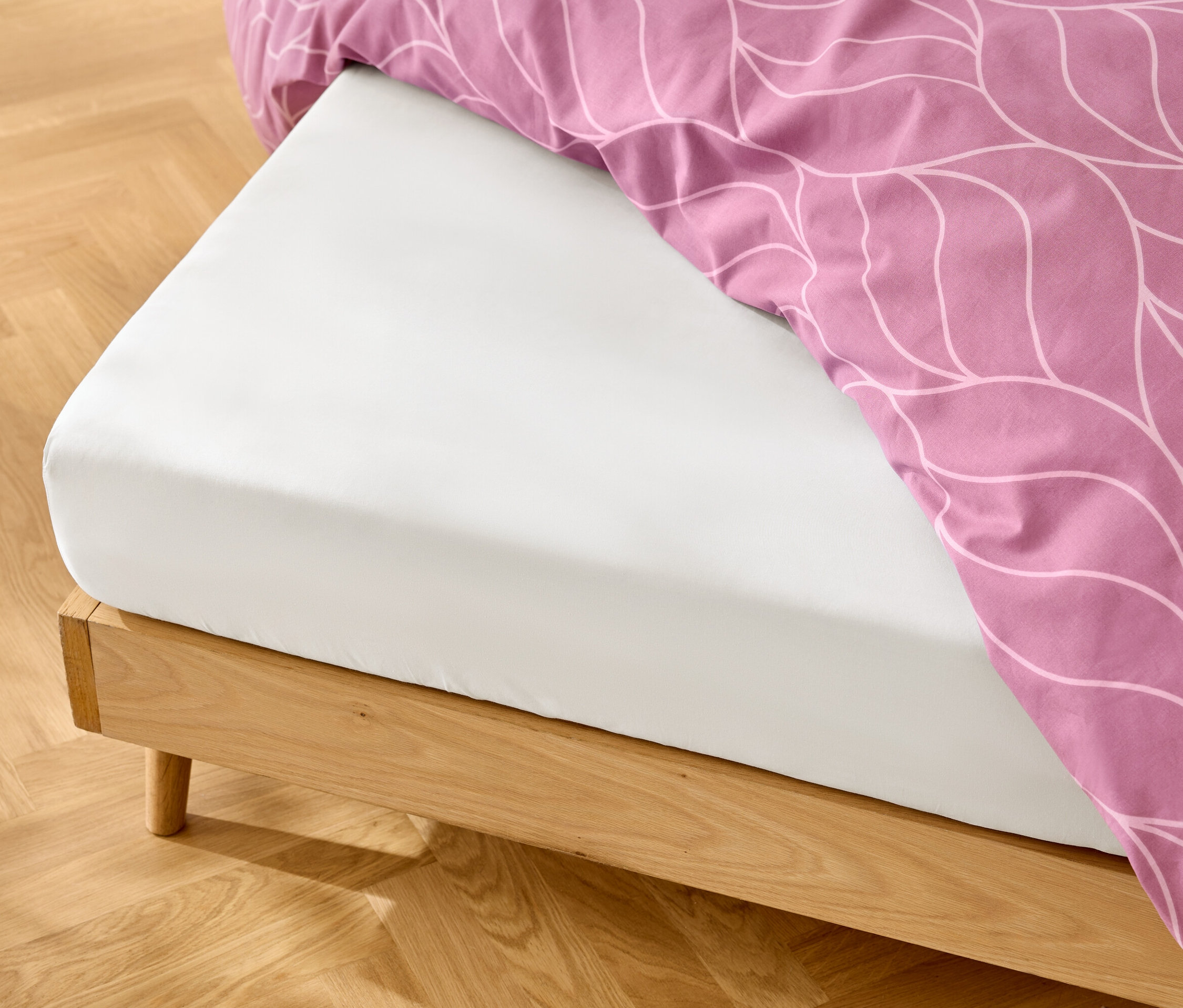 Bílé prostěradlo na posteli s dřevěným rámem a růžové povlečení s bílým vzorem.