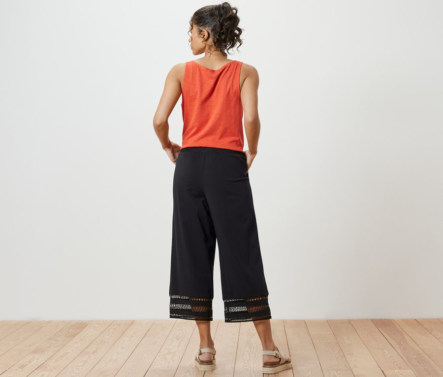 Žena stojí zády a má na sobě oranžový top a černé culotte kalhoty s výšivkou.
