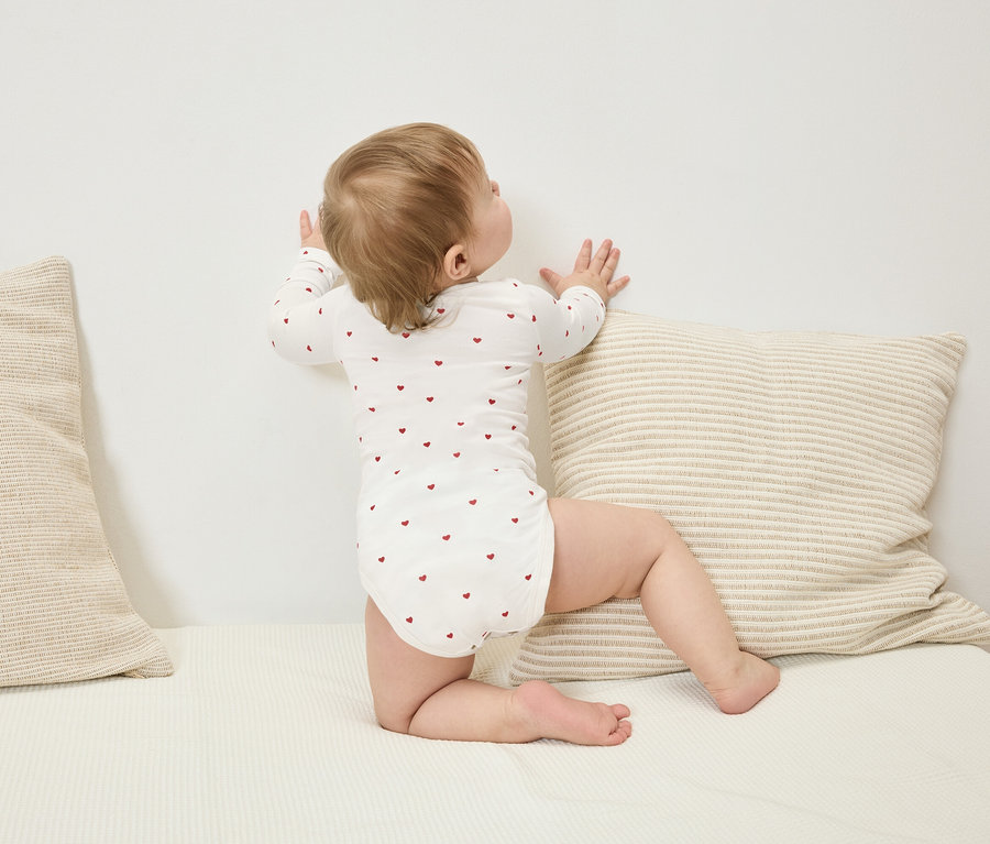 Batole v body s dlouhým rukávem s červenými srdíčky stojí u zdi vedle pruhovaného polštáře.