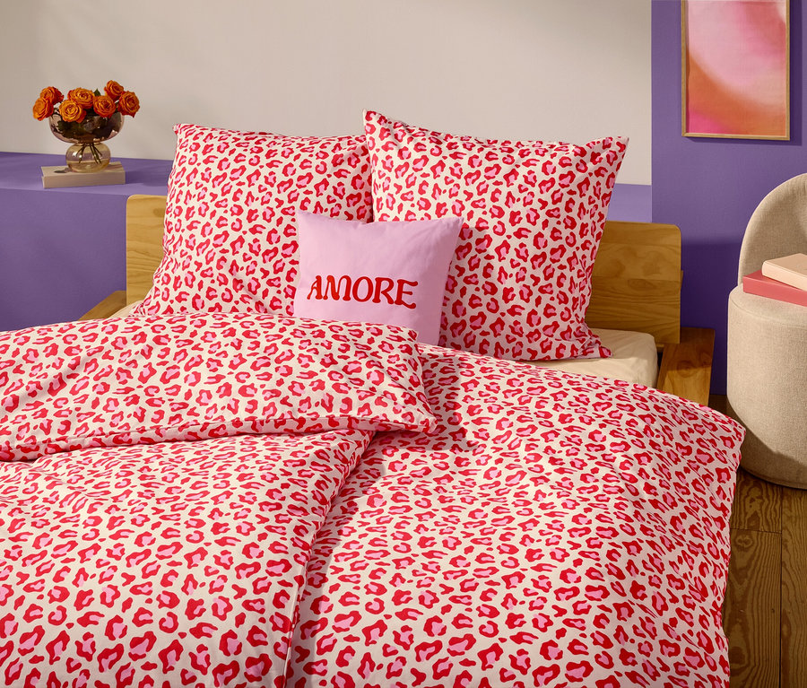 Detailní záběr na postel s červeno-bílým ložním prádlem s leopardím vzorem a polštáři. Levandulový polštář s nápisem "AMORE" je umístěn před dvěma polštáři.