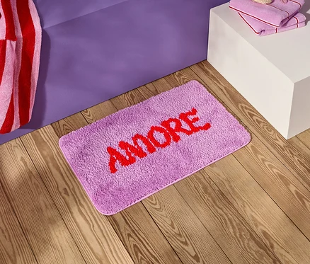 Na dřevěné podlaze leží fialový koupelnový kobereček s červeným nápisem "AMORE" vedle fialového nočního stolku.