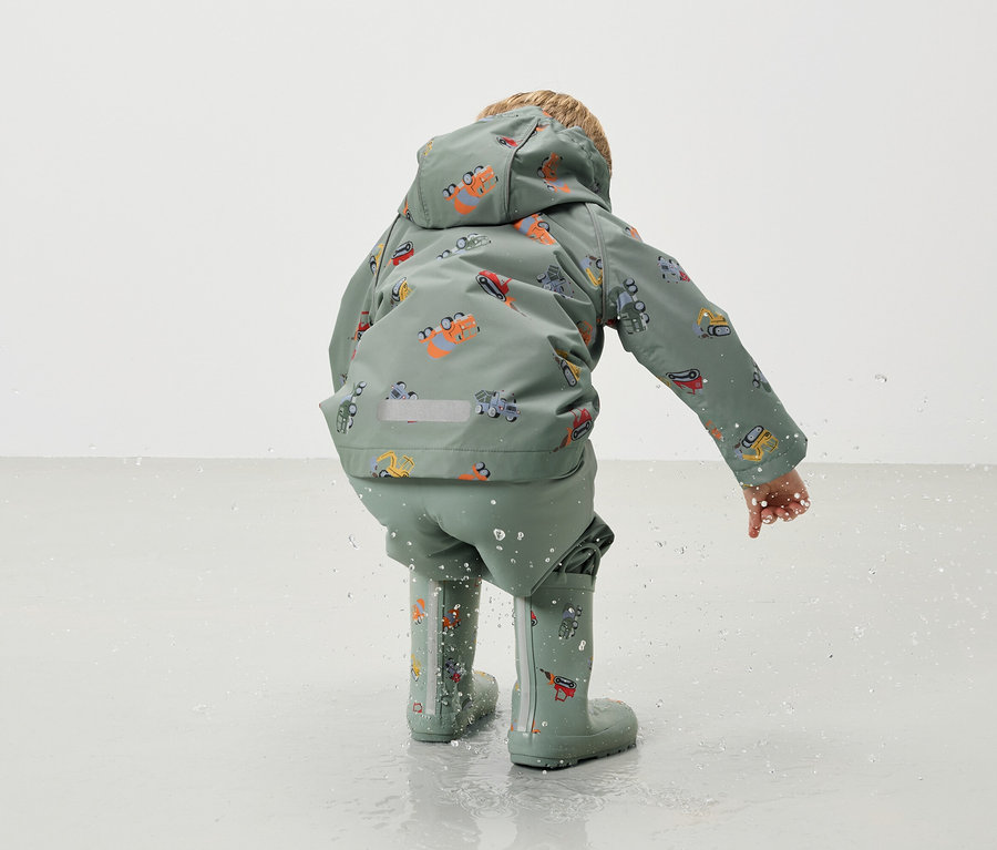 Dítě zezadu, má na sobě zelenou bundu do deště a kalhoty do deště s motivem bagru a odpovídající gumáky, přičemž si hraje ve vodě.