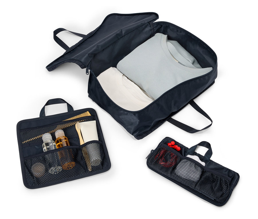 Tři otevřené, tmavě modré cestovní tašky s držadly a zipy uspořádané na bílém pozadí. Tašky obsahují oblečení, kosmetiku a doplňky.