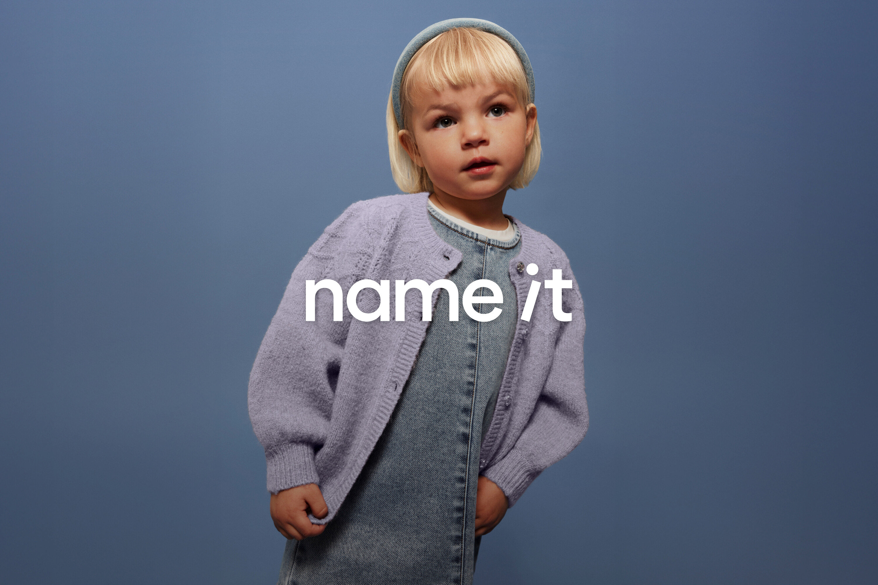 Malé dítě s blond vlasy a modrou čelenkou má na sobě džínové lacláče a fialový kardigan na modrém pozadí.