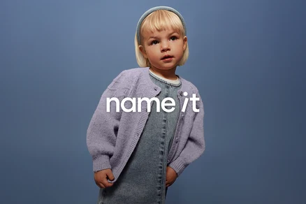 Malé dítě s blond vlasy a modrou čelenkou má na sobě džínové lacláče a fialový kardigan na modrém pozadí.