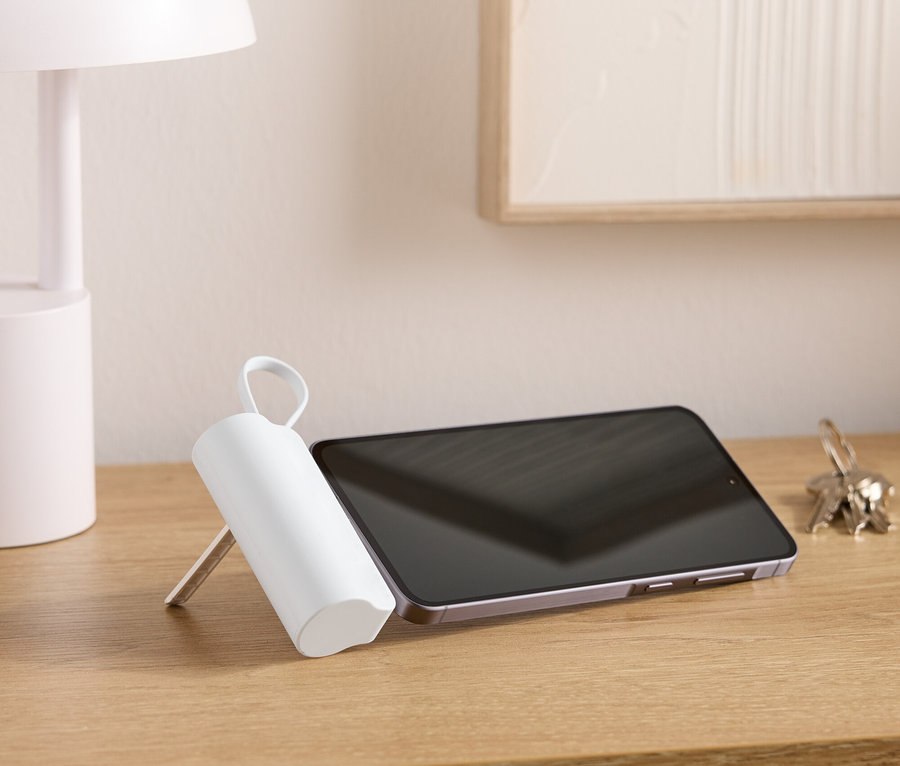 Na dřevěném stole stojí lampa, tablet, svazek klíčů a bílý válcovitý předmět s rukojetí.