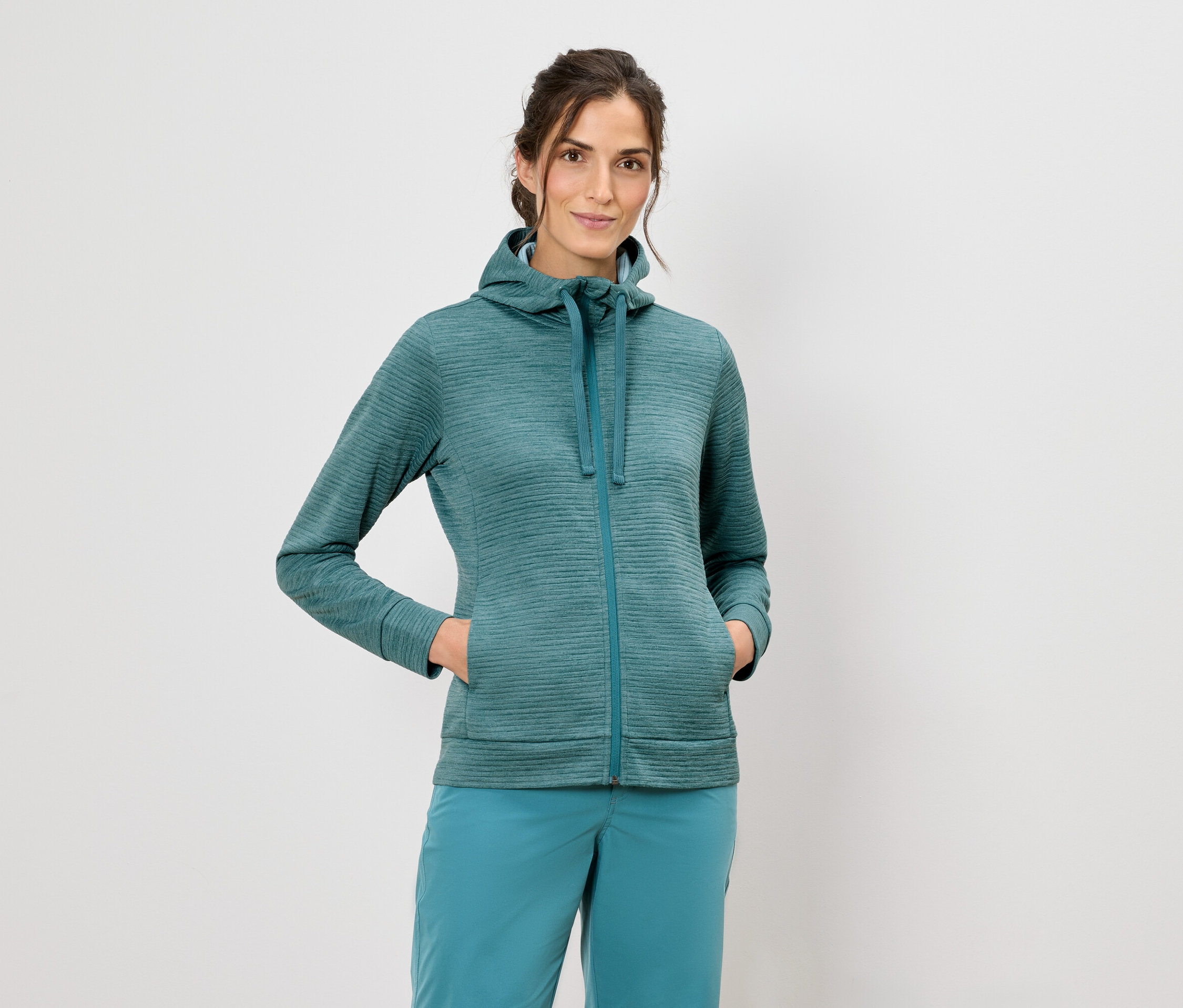 Žena v mikině s kapucí a modrých kalhotách stojí na bílém pozadí.