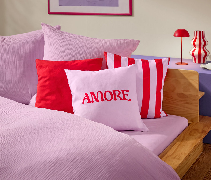 Detailní záběr postele s růžovým ložním prádlem a polštáři. Na jednom z polštářů je červeným písmem napsáno slovo AMORE.