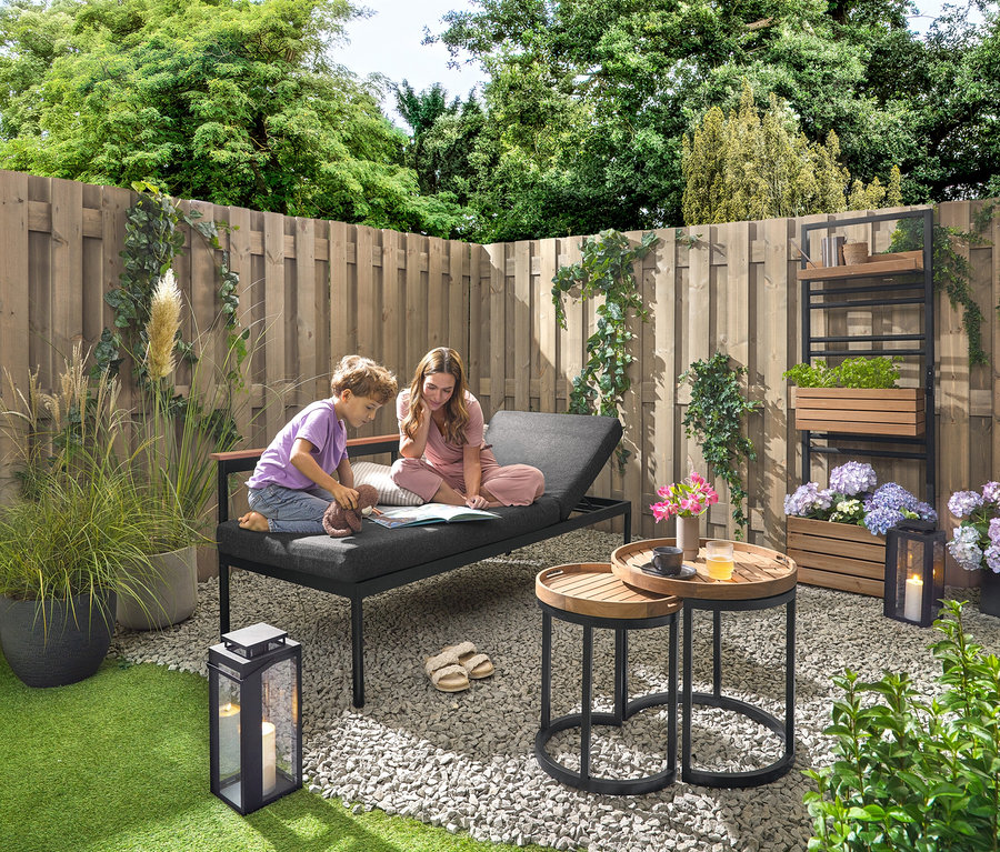 Dítě a žena sedí na zahradní lavici 2 v 1 ‒ lehátku »Elin« a čtou si knihu. V pozadí je zelená stěna s flexibilně zavěsitelnými policemi »Elin«. Vpravo stojí venkovní lucerna se svíčkou. K dispozici jsou také 2 zahradní odkládací stolky s odnímatelnými podnosy, průměr cca 40 cm a 50 cm, a 2 venkovní polštáře s lněným omakem.