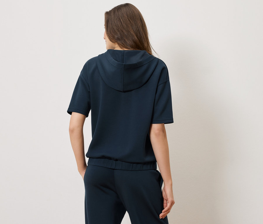 Pohled zezadu na ženu v tmavě modré mikině s kapucí a krátkým rukávem a odpovídajících kalhotách.